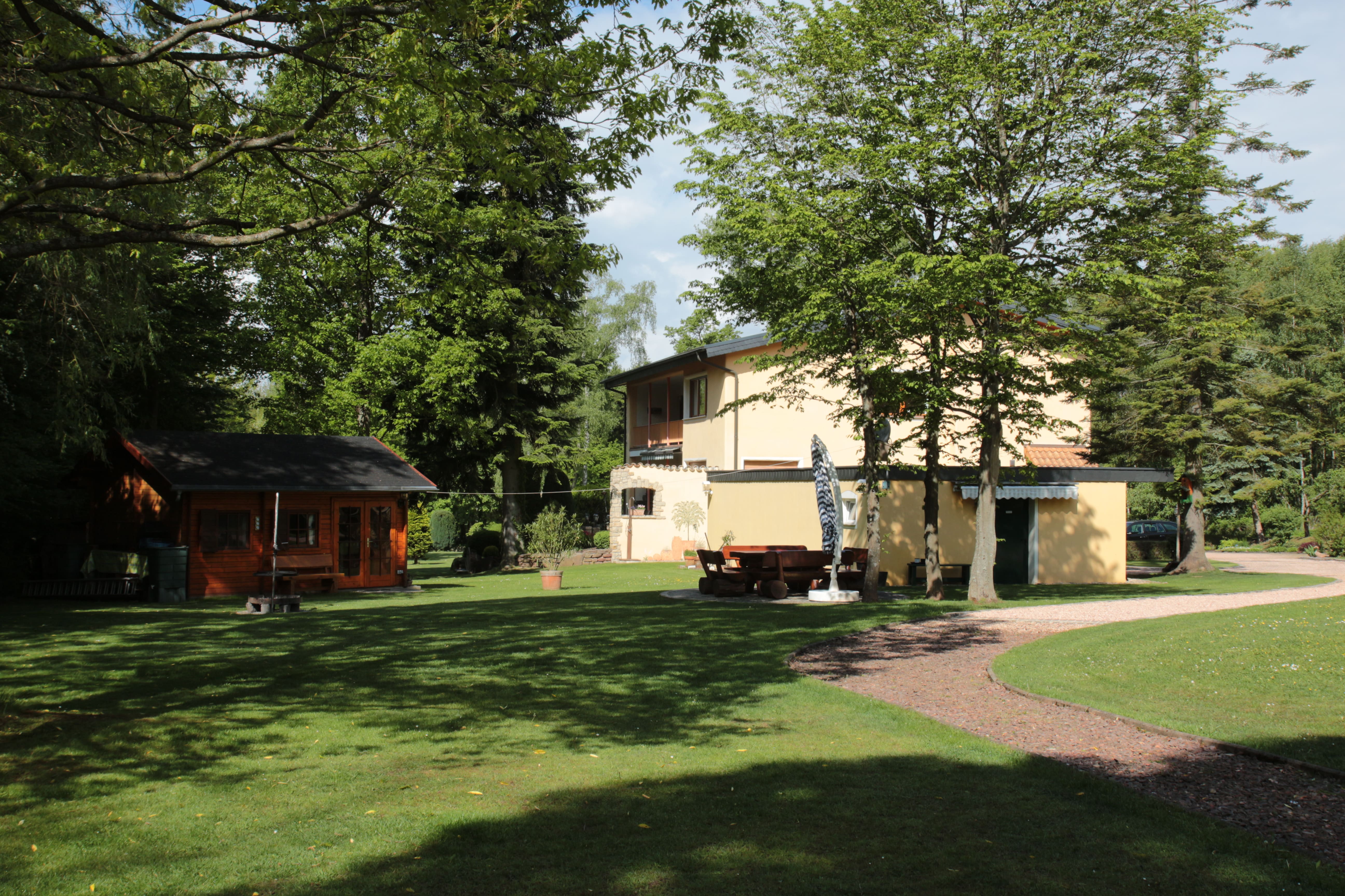 Rückseite der Pension Rehwinkel in Nalbach, umgeben von großzügiger Grünfläche, mit einem kleinen Holzhaus und einem Weg, eingebettet in die Natur.