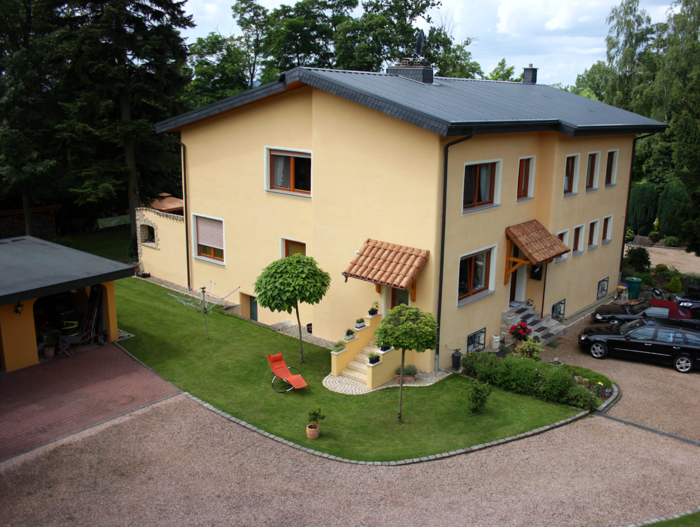 Rückansicht der Pension Rehwinkel in Nalbach, mit gepflegtem Gartenbereich, gepflasterter Einfahrt, Parkmöglichkeiten und einer gemütlichen Sitzecke mit Liegestuhl.