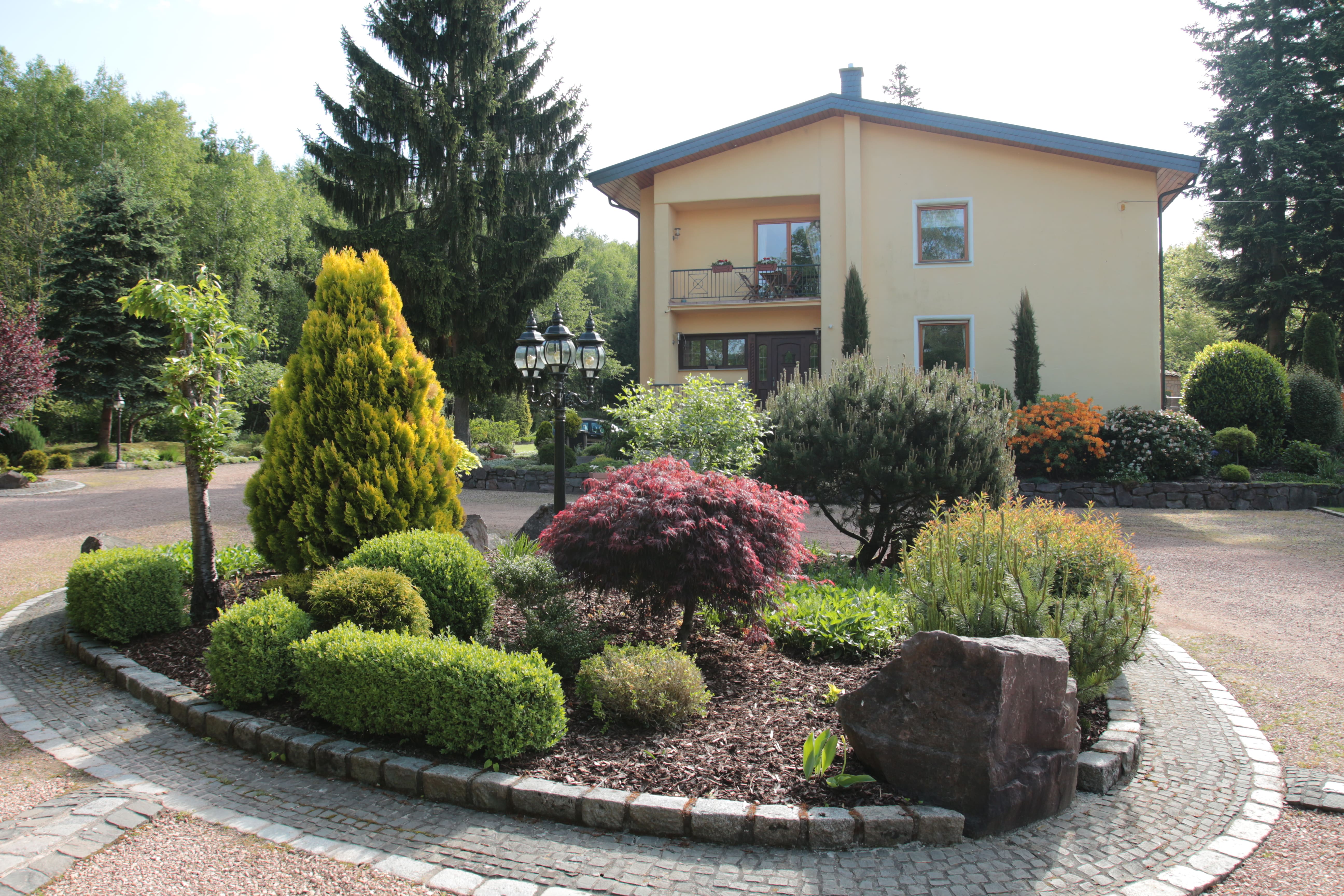 Eingangsbereich der Pension Rehwinkel in Nalbach, mit gepflegtem Garten, Steinweg und einem zweistöckigen gelben Gebäude im Hintergrund, umgeben von Bäumen und Natur.
