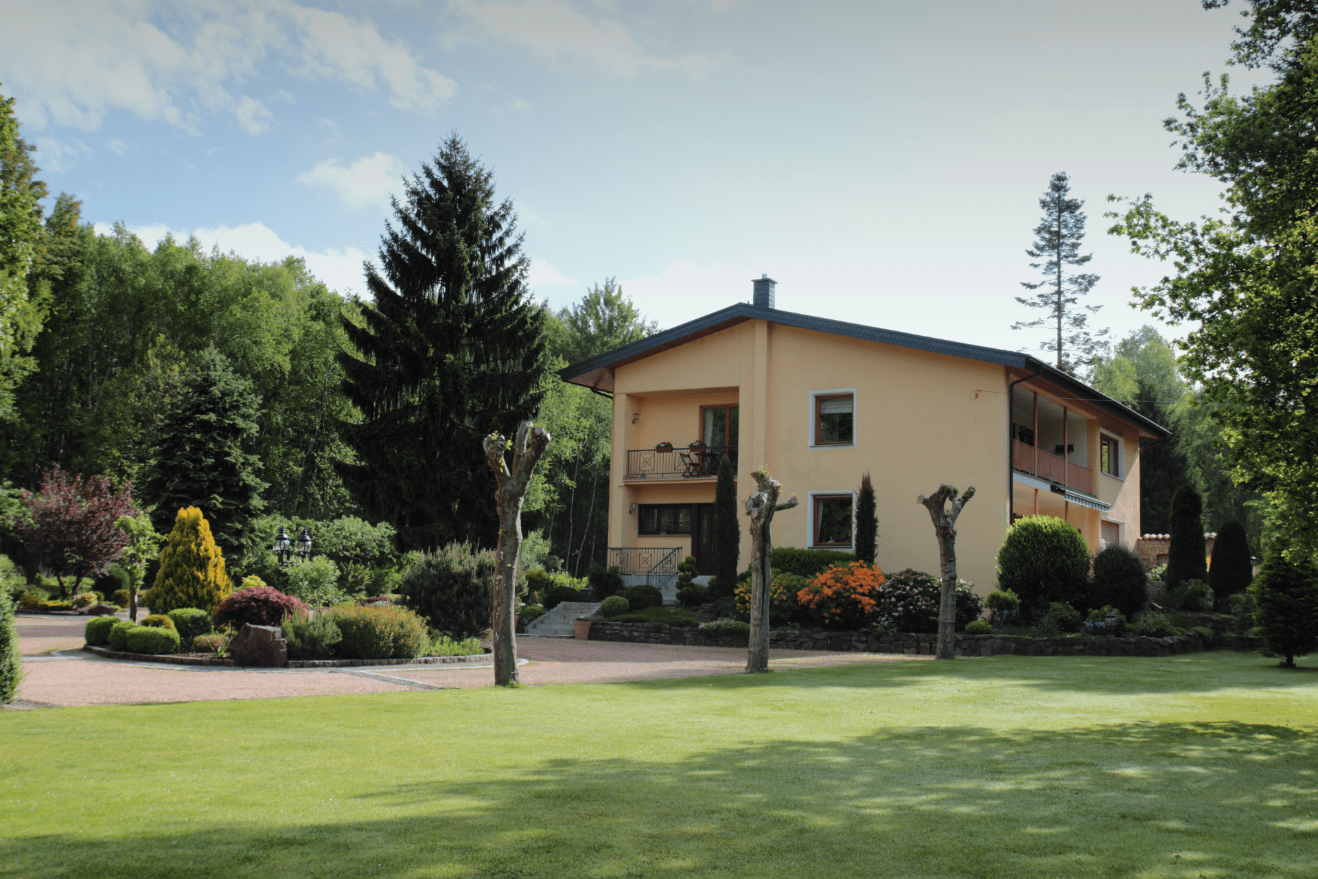 Vorderansicht der Pension Rehwinkel in Nalbach, umgeben von gepflegtem Rasen und hohen Bäumen, mit einer einladenden Gartenanlage.